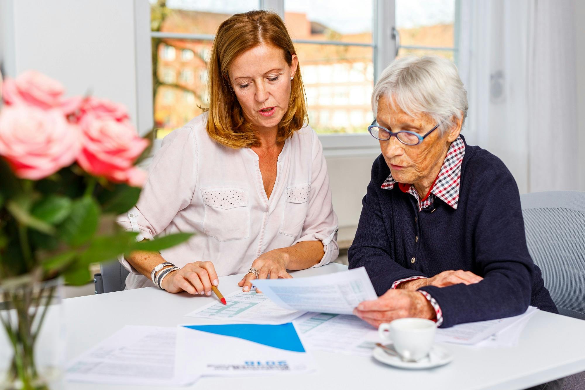 Jüngere Frau hilft älterer Dame bei der Dokumentenprüfung am Tisch mit Unterlagen und Kaffeetasse.