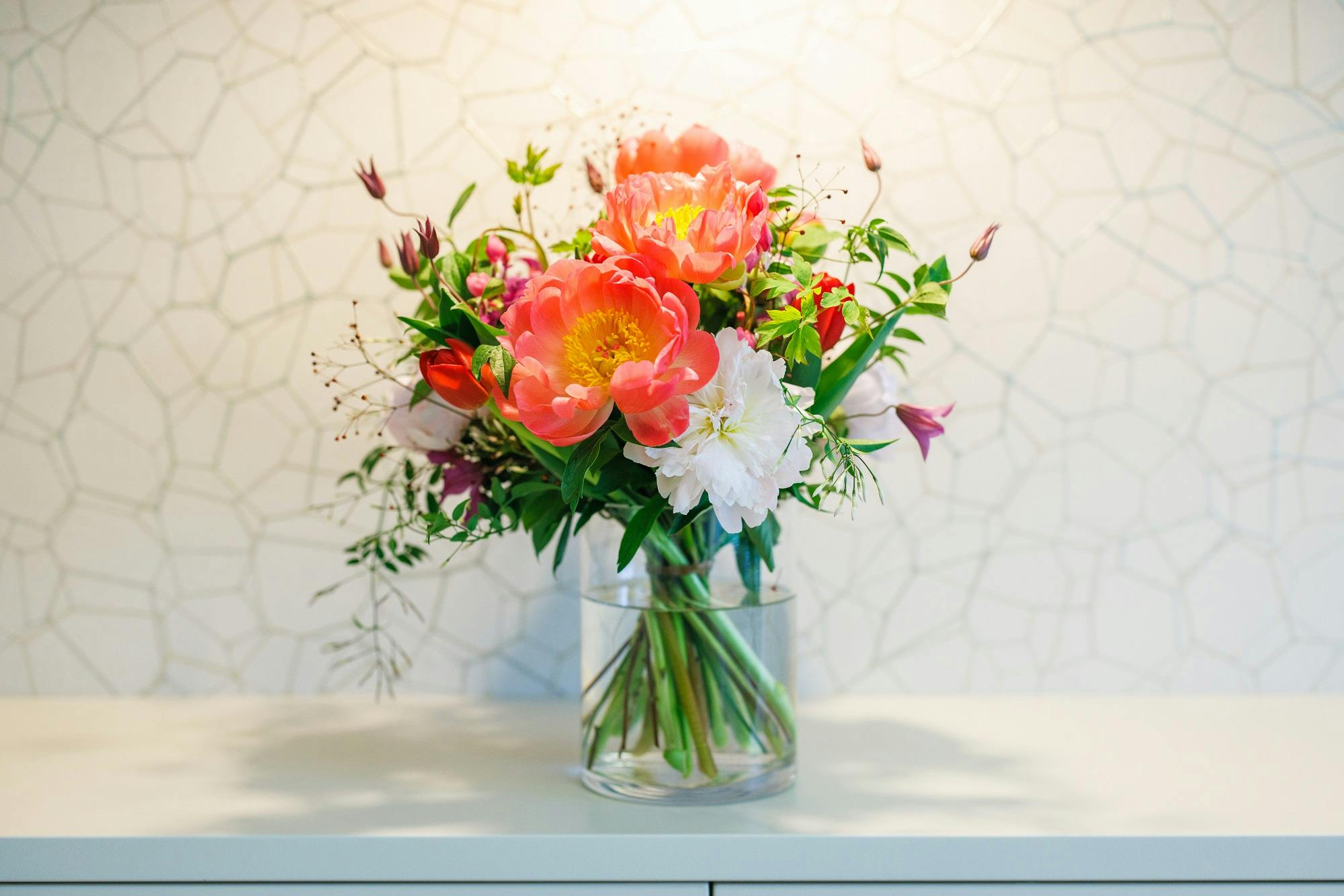 Bunter Blumenstrauß in einer Glasvase auf einem weißen Tisch vor Mosaik-Hintergrund.