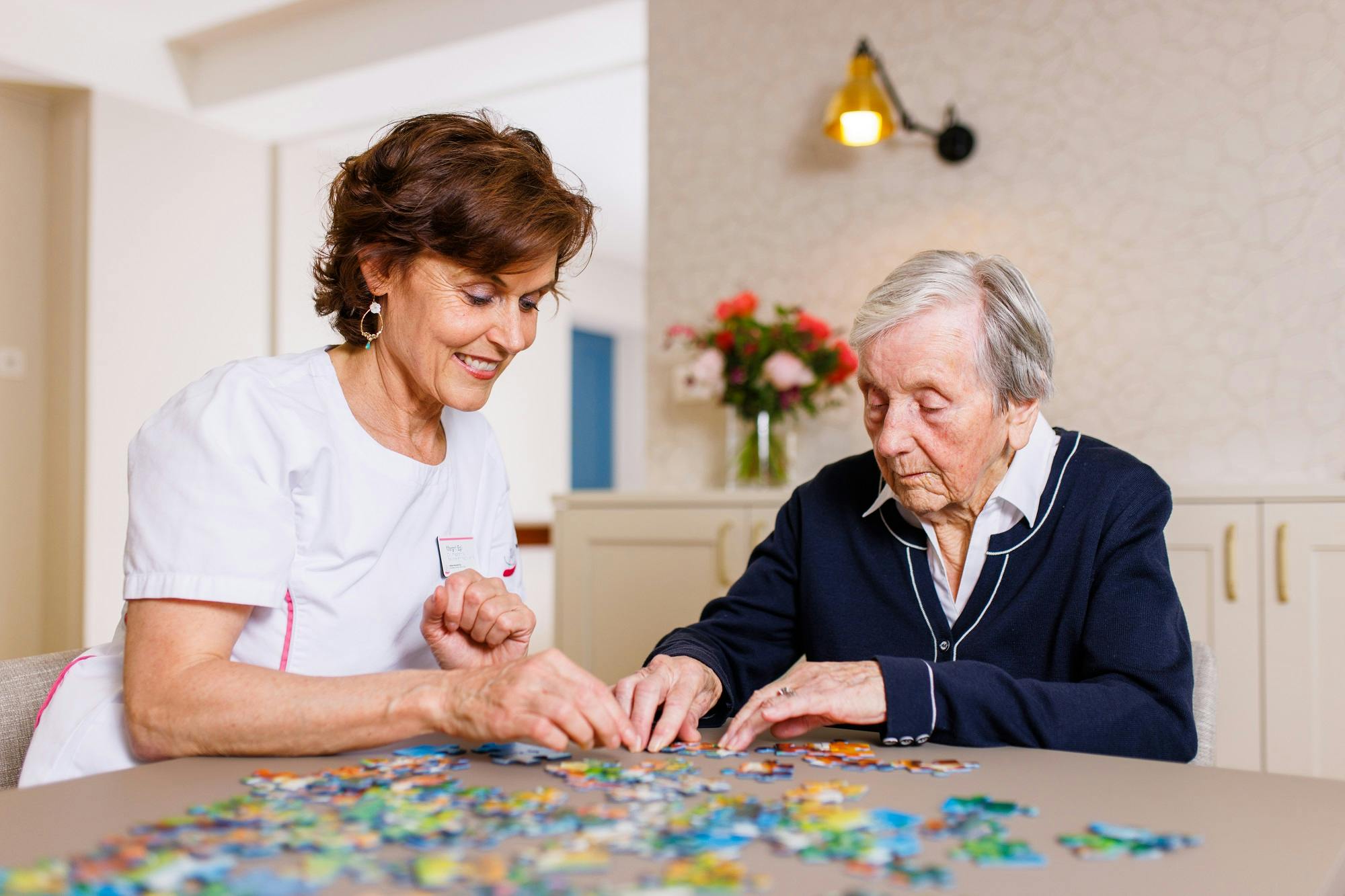 Pflegekraft unterstützt Senior beim Puzzeln im Wohnzimmer.